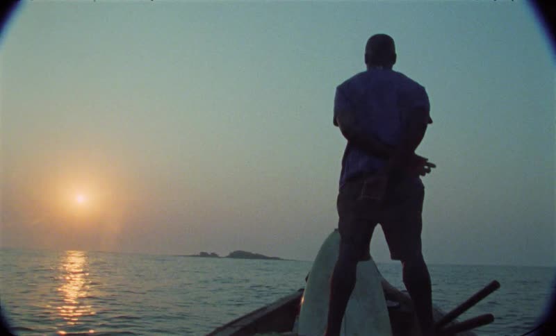 A man stands at the prow of a moving boat while looking out over the sea at dusk