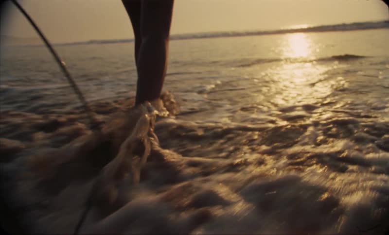 Following a young woman entering the ocean with a surfboard at evening