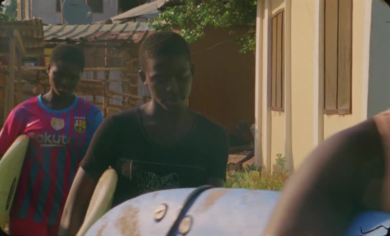 A group of women carry surfboards while walking through a village during the day