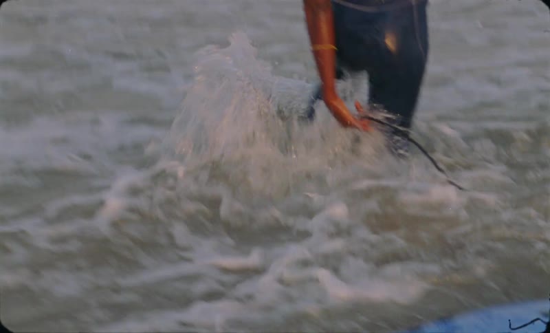 A smiling woman rides a surfboard towards the beach before hopping off in the shallow water