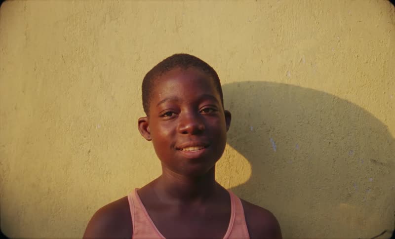 African boy smiling at the camera while standing near a yellow stucco wall then gazes in the distance