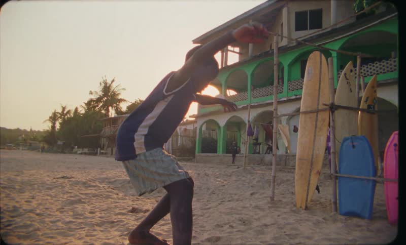 Youth practices for surfing by balancing on wooden board near cluster of surfboards as sun shines in red tinted sky