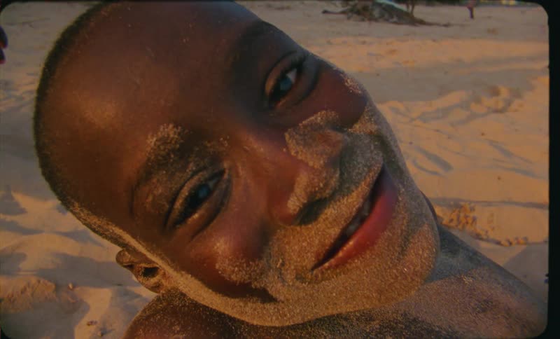 A young boy is lays buried on the beach with sand on his face and smiles at the camera