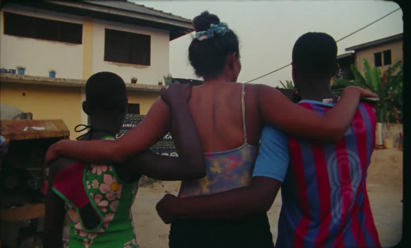 A group of women walk together through a village with their arms around one another