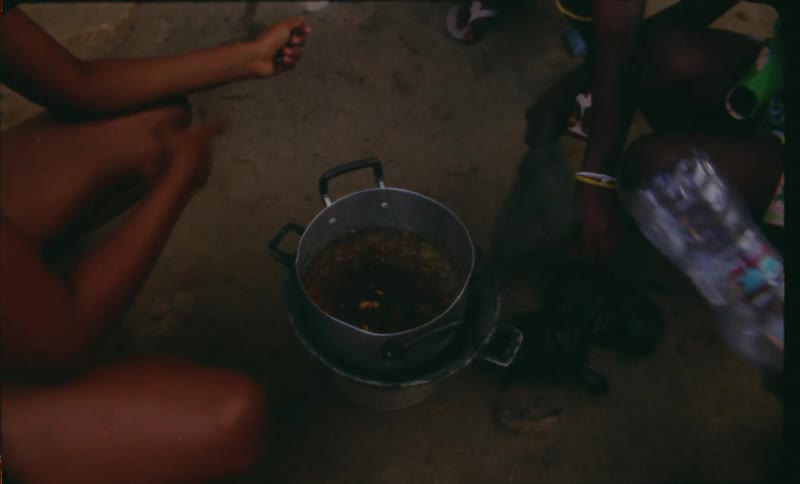 A group of about 4 people including kids sit around a pot of soup cooking while one child fans it with a hand fan