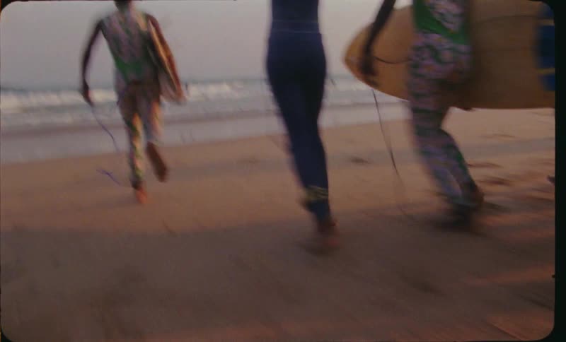 A group of young black girls run down the beach toward the water while carrying surfboards