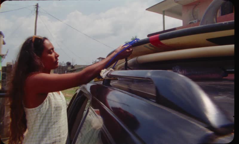 A young woman uses cable ties to strap a surfboard to the top of her car