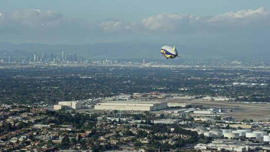 Aerial footage of the Goodyear Blimp approaching an airport with the Downtown LA skyline in the background in Long Beach California during the day