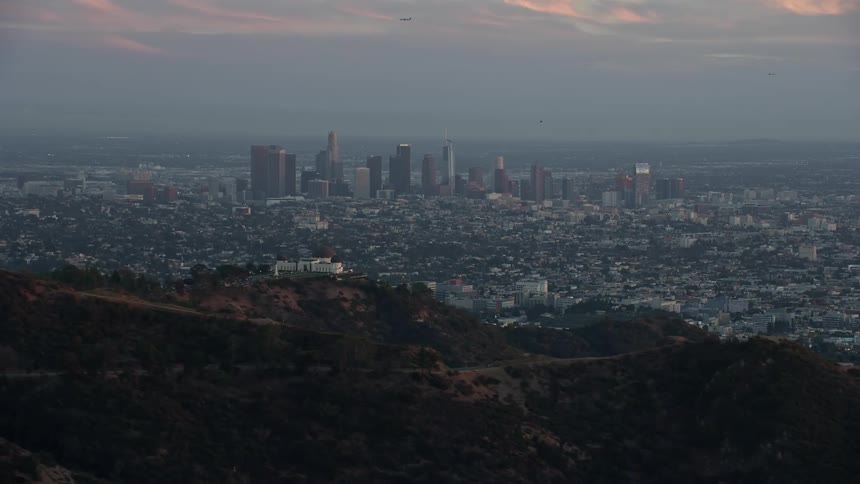 An epic aerial footage showcasing the Downtown Los Angeles skyline at twilight ending with a reveal of the iconic Griffith Observatory shot during summer with partly cloudy skies in the background