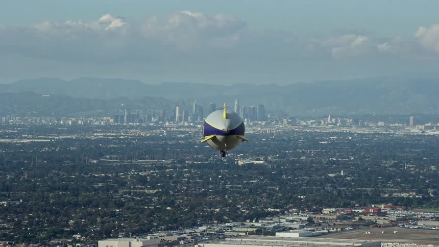 A Goodyear Blimp flies towards the Downtown Los Angeles skyline from Long Beach Airport in California with partly cloudy skies and cityscape shot with a Shotover F1 camera during daytime aerials of Los Angeles California during summer