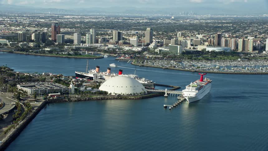 Aerial footage of a cruise ship and the historic Queen Mary docked in Downtown Long Beach CA on a partly cloudy day during the spring or summer season