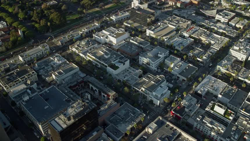 A stunning aerial view of Beverly Hills California shows a flyover of office buildings and shops while tilting down to Rodeo Drive and the shopping centers during spring or summer
