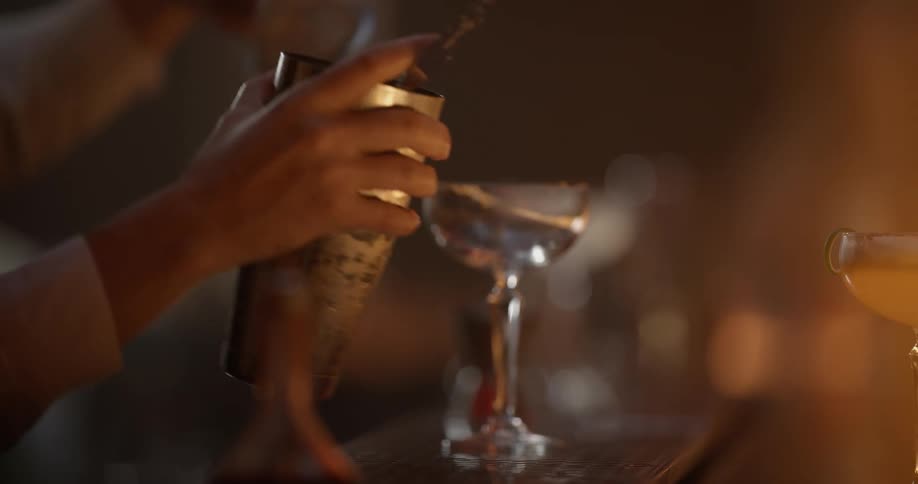 A mixologist pours a drink from a cocktail shaker at a bar
