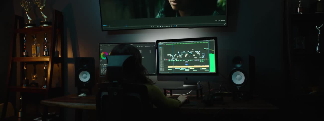 A large television above two computer monitors in front of a woman sitting at the desk