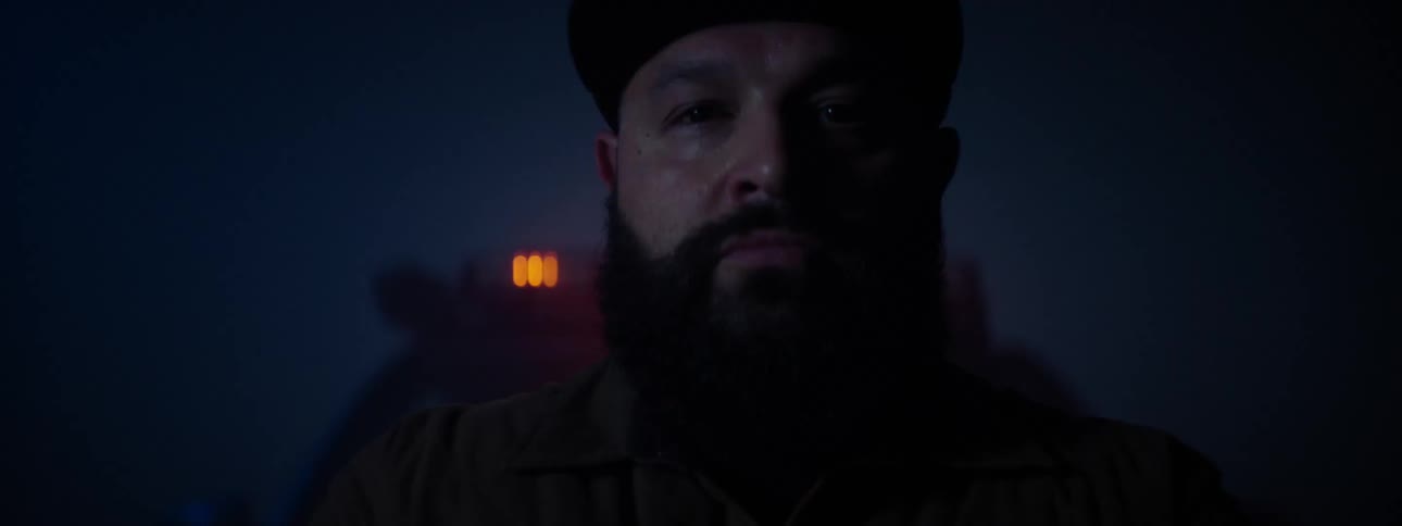 A man with a beard and wearing a hat is standing in front of an automobile and bright lights come on behind him