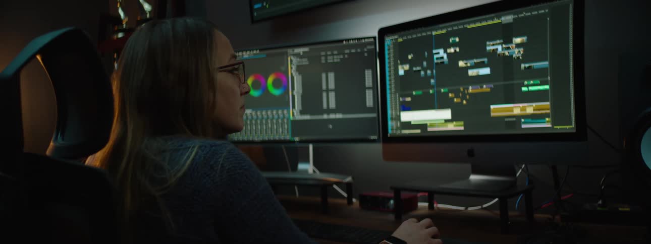 A woman is sitting at a desk working on a computer as she looks at the monitors