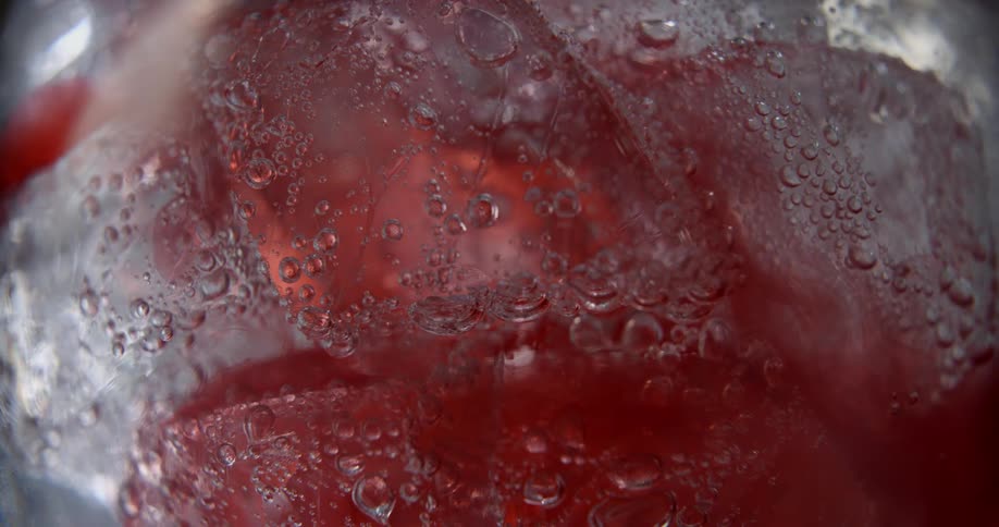 An extreme close up of ice in the bottom of a glass with a red liquid being added to the clear substance in the glass with ice