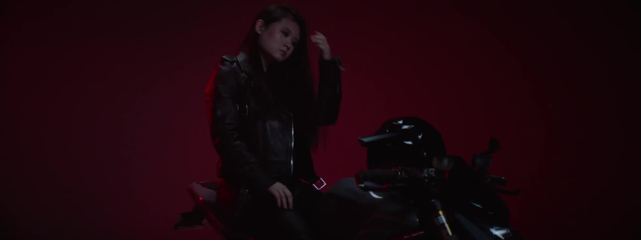 A woman wearing a black leather jacket carrying a helmet walks over to a motorcycle and gets on it setting the helmet on it and grabbing the handle bars