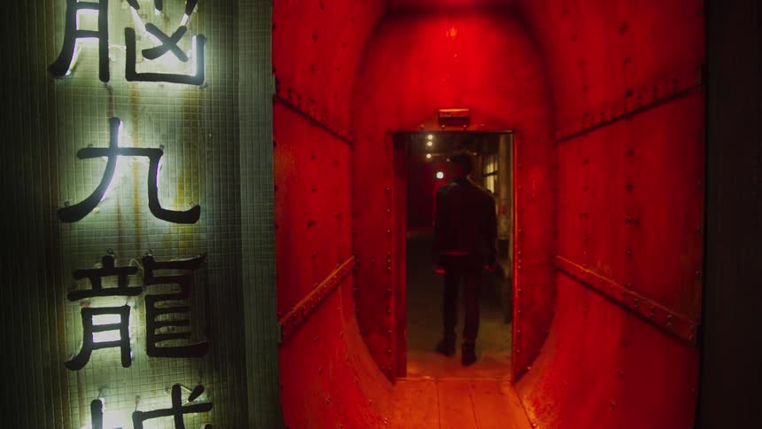 A man standing outside of a building reading a sign then turning and walking down a red lit hallway through a door