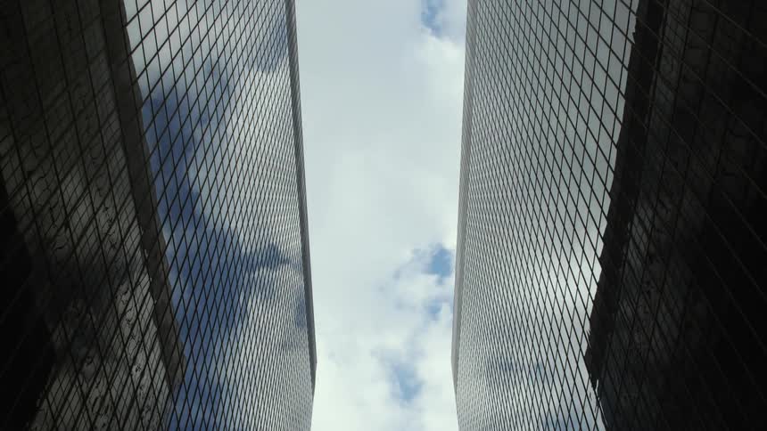 A straight up view between two large tall buildings covered in windows with a cloudy blue sky between them