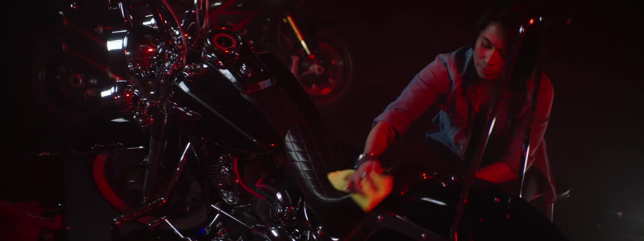 A woman knelt down using a rag to wipe down a motorcycle seat with a red light cast on her