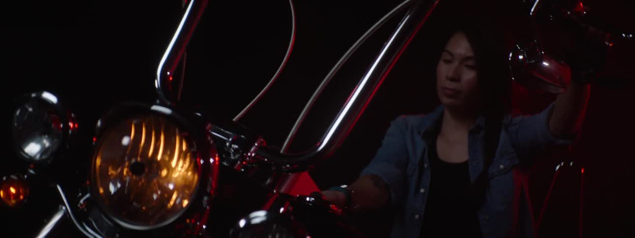 Woman with blue shirt and black top gets on motorcycle in dark room