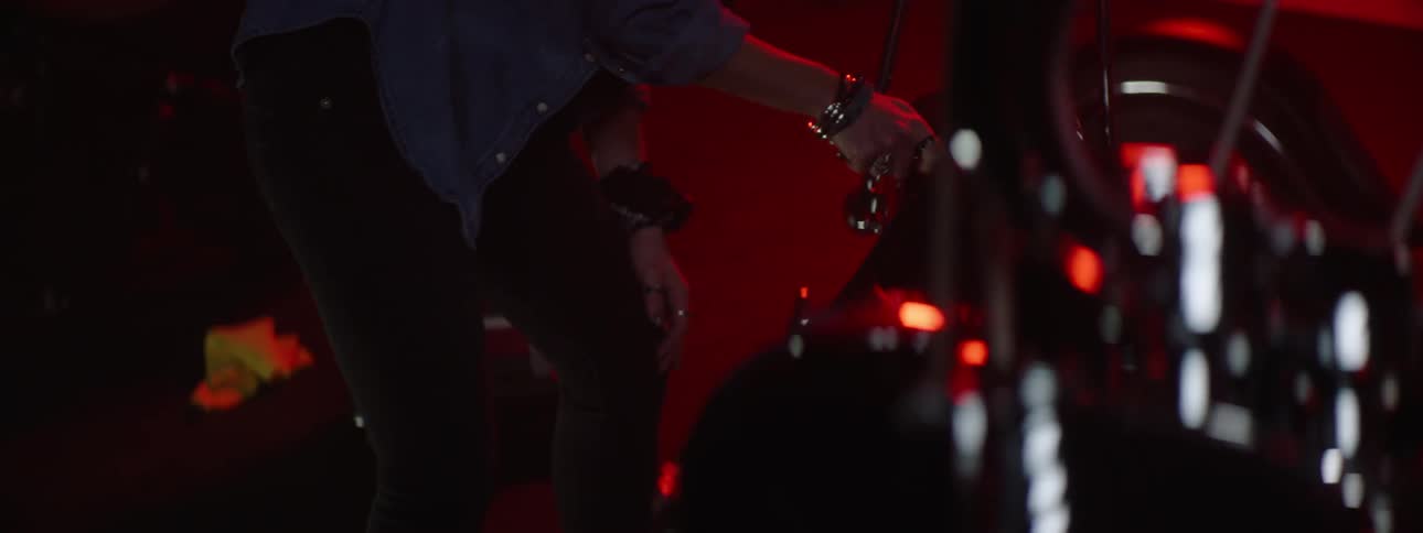 A man walking through a garage lit by red light as he walks over to a motorcycle and uses a tool on it