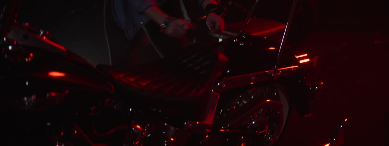 A woman walking up to a motorcycle and uses a tool to take the seat off in a red lit area