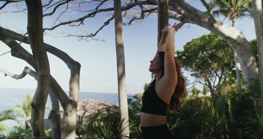 A woman standing outdoors stretching near trees with the ocean in the background