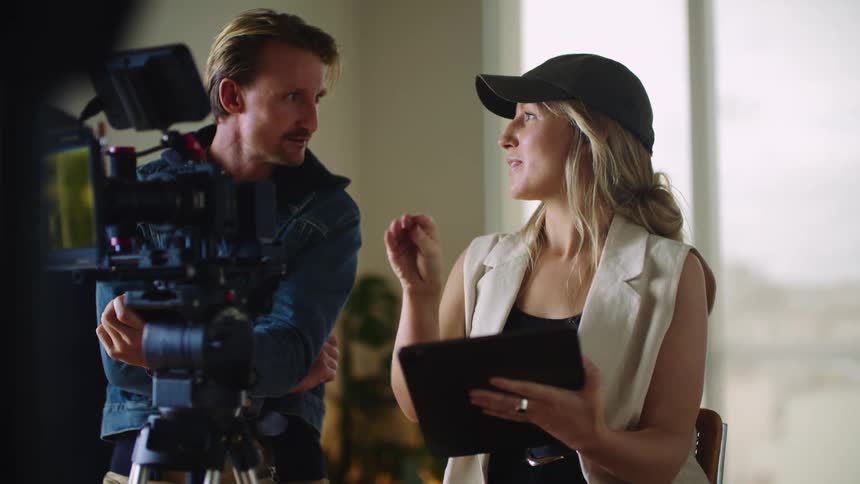 Blonde woman with black cap top and white vest talks to man in blue jacket behind the movie camera