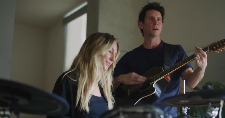 A woman with long blond hair is playing the drums as a man is standing next to her is playing an acoustic guitar
