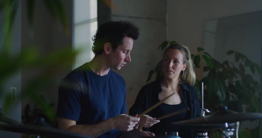 Woman with ponytail black jacket and top shows man how to play drums