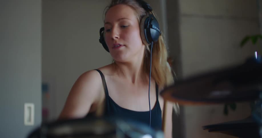 Blonde woman with ponytail black top and headphones play drums in room