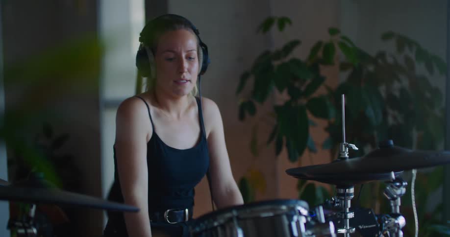 A woman wearing headphones is holding drumsticks as she plays the drums in a room with plants near the wall