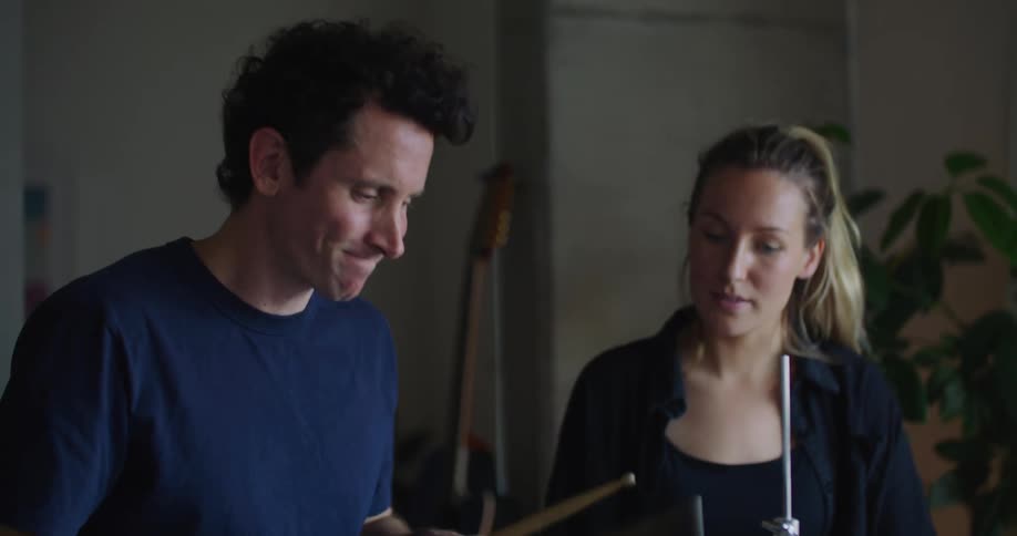 Blonde woman with ponytail and black shirt shows man how to play drums