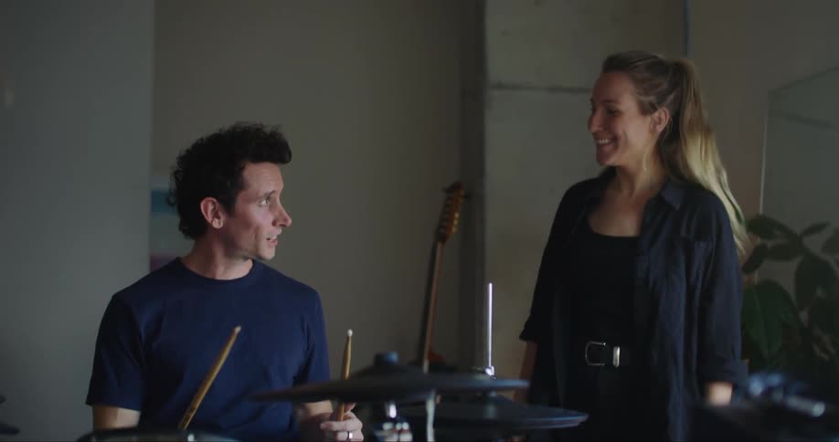 A man is holding drums sticks as he plays the drums and the woman standing next to him is smiling and pats him on the shoulder as she cheers him on