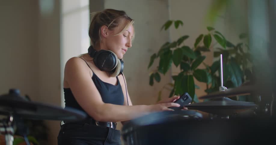 A woman playing a drum set with headphones on then picking up her cellphone to talk on it