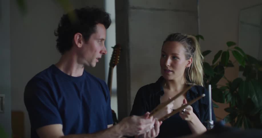 Woman with ponytail black jacket and shirt shows man how to play drums