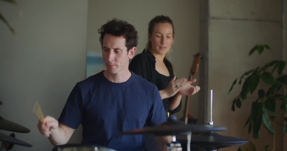 Woman with ponytail and black shirt clasping hands while man playing drums in room