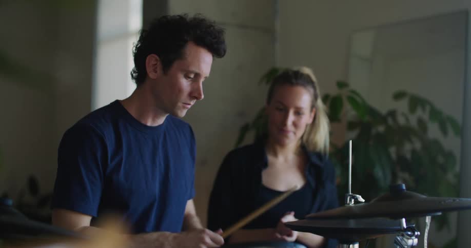 Blonde woman with ponytail and black shirt shows man how to play drums
