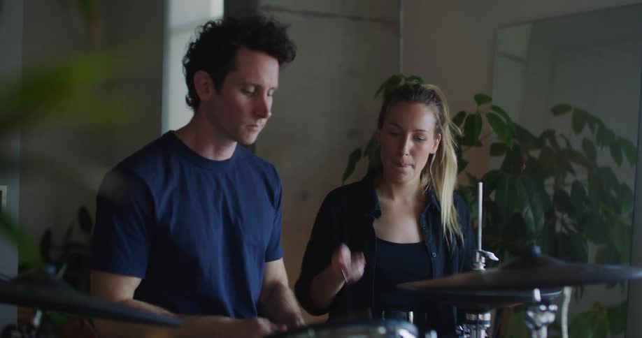 Blonde woman with ponytail and black shirt shows man how to play drums
