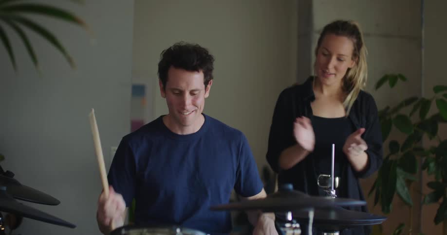 Man playing ending fill on 5piece drum kit while woman claps along behind him