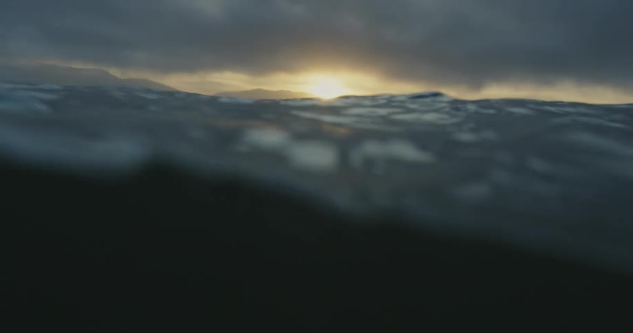 Sun shines over mountains on coast under overcast sky and is reflected by ocean waves