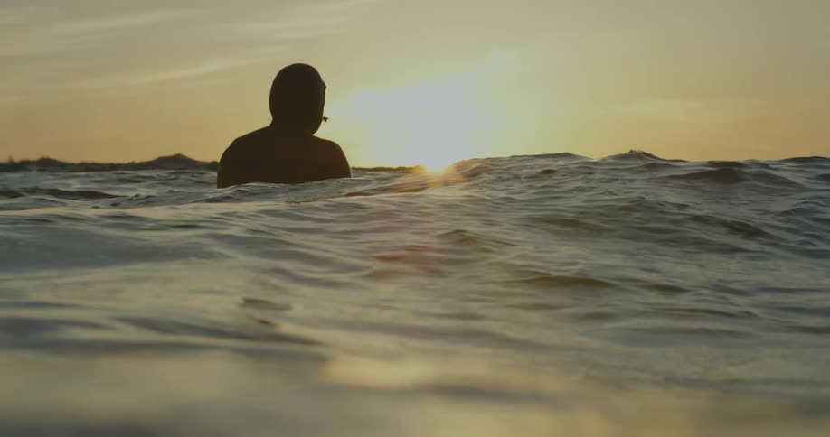 A man is standing in the water as waves move and reflect light with the sun shining brightly on the horizon
