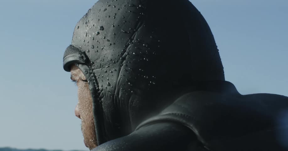 A man wearing a wetsuit is lying on his surfboard as he paddles with his arms across the water