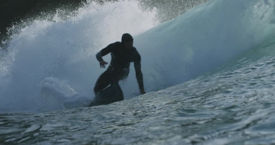 Surfer wearing wet suit rides board along crashing wave passing promontory