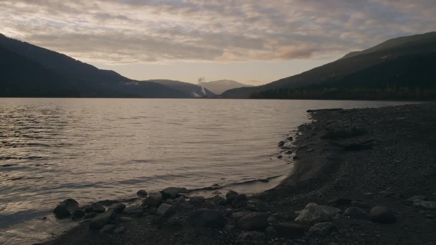 Waves roll onto shore across lake from smoke rising between mountains on misty horizon
