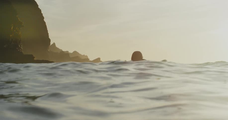 Man in wetsuit swims on surfboard in water at sunset