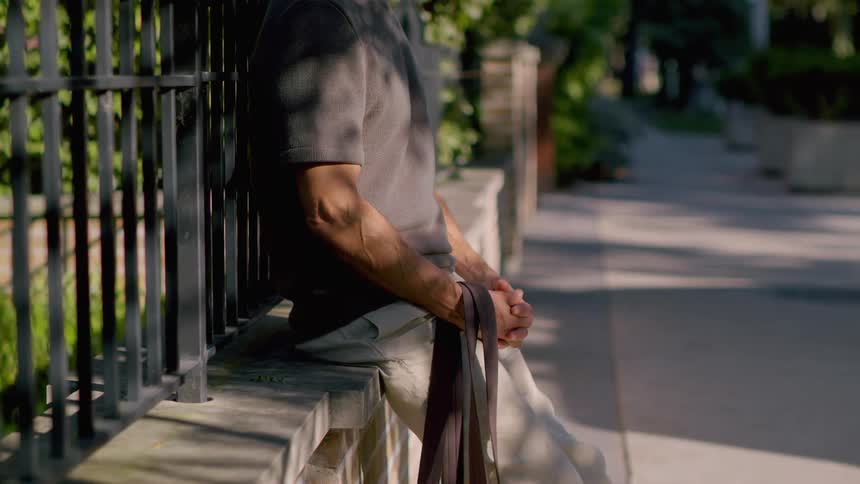 A mans hands are clasped in front of him as he leans against a fence wall in a park