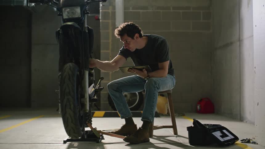 A man is sitting on a stool reaching into a tool box as he holds a tablet computer and works on a motorcycle
